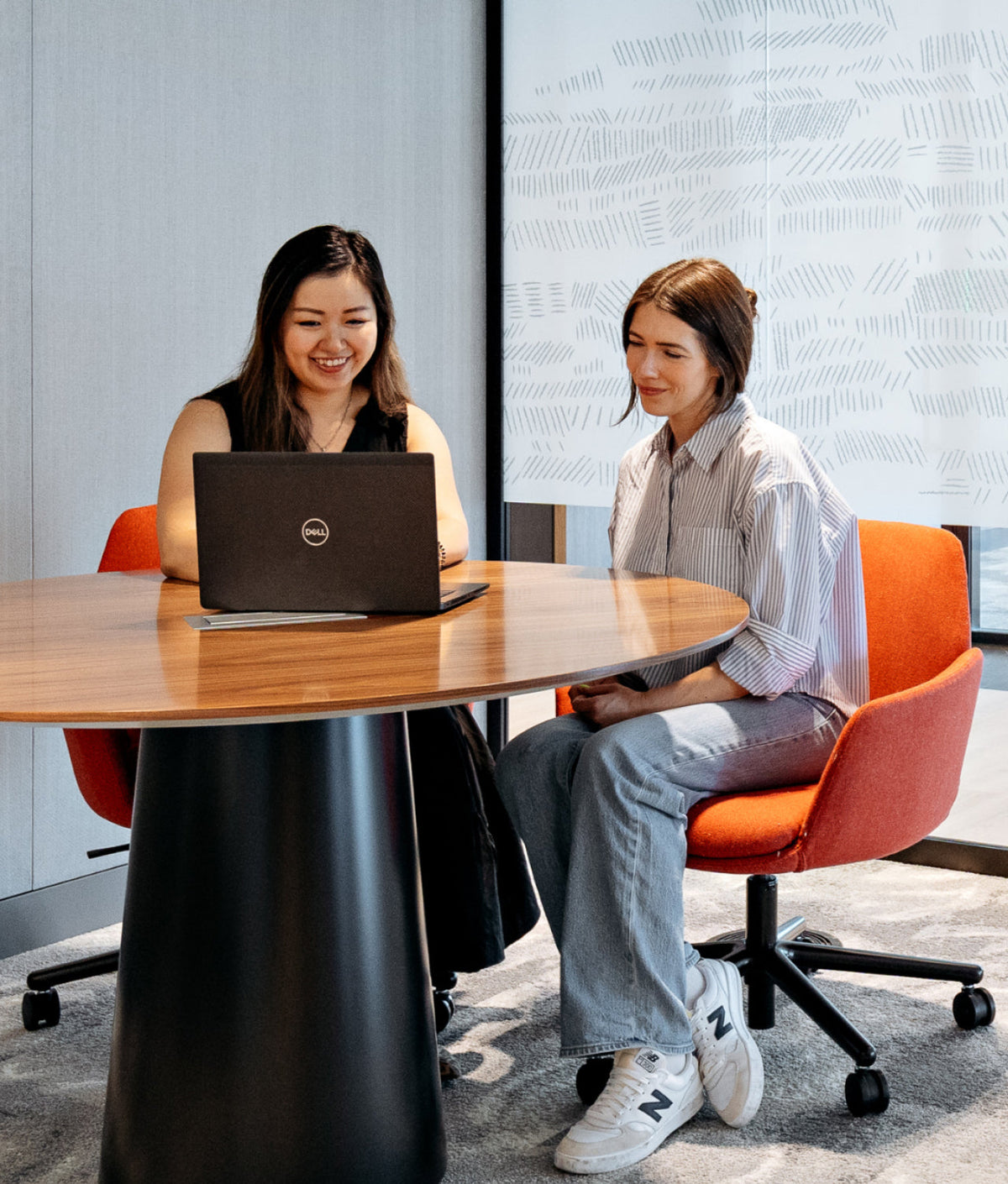 Two women collaborating at a round wooden table in a modern office setting, with one using a laptop and the other sitting on an orange chair.