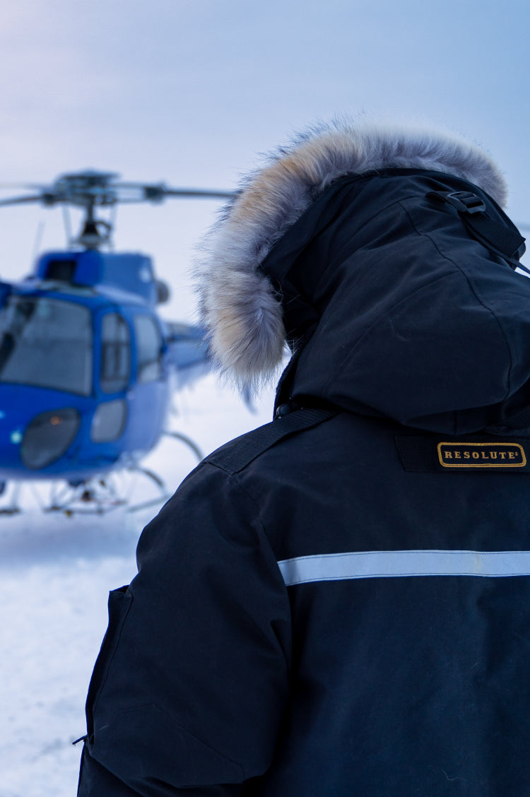 A person wearing a dark navy Resolute winter parka with a fur-trimmed hood stands in a snowy outdoor setting with a blue helicopter in the background.