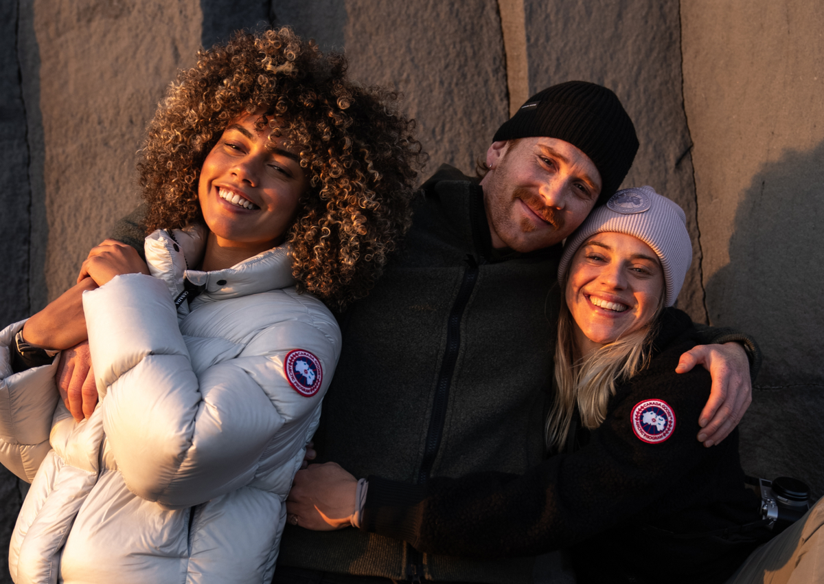 Three people hugging outdoors, each wearing a Canada Goose puffer jacket with the brand's logo patch visible on the sleeve.